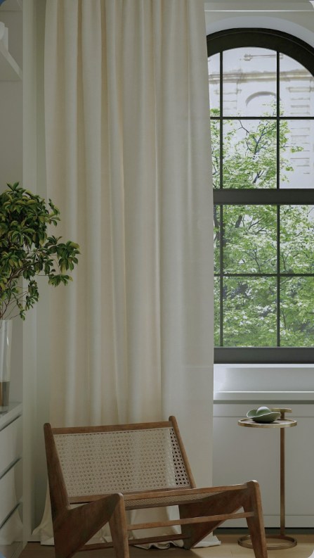 A cane chair beside cream curtains and an arched window